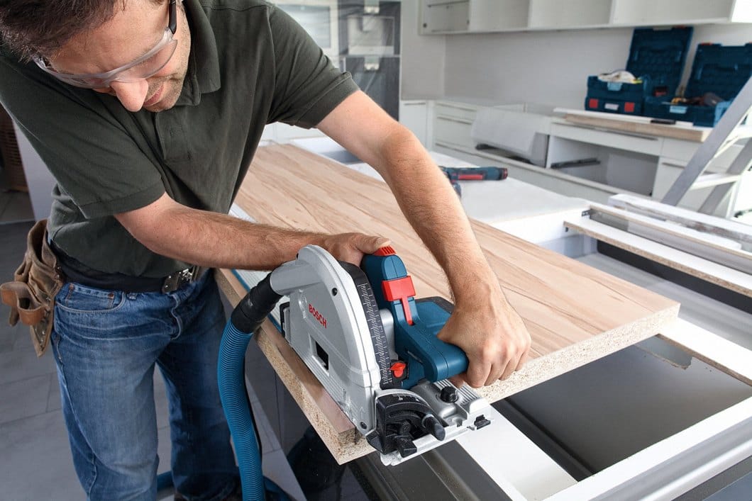 Individual cutting laminated wood with Bosch track saw connected to vacuum hose; toolboxes and boards in background.