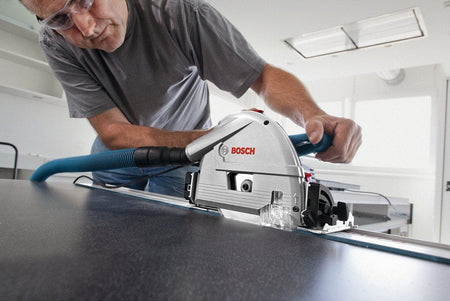 Person using Bosch track saw guided along metal track to cut wood on workbench; safety glasses worn in workshop.