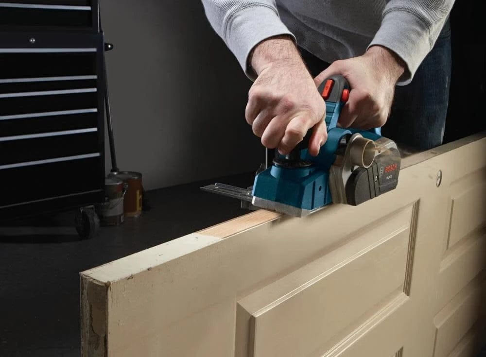 Bosch hand planer shaving top edge of wooden door; tool guided with both hands for precision adjustment.