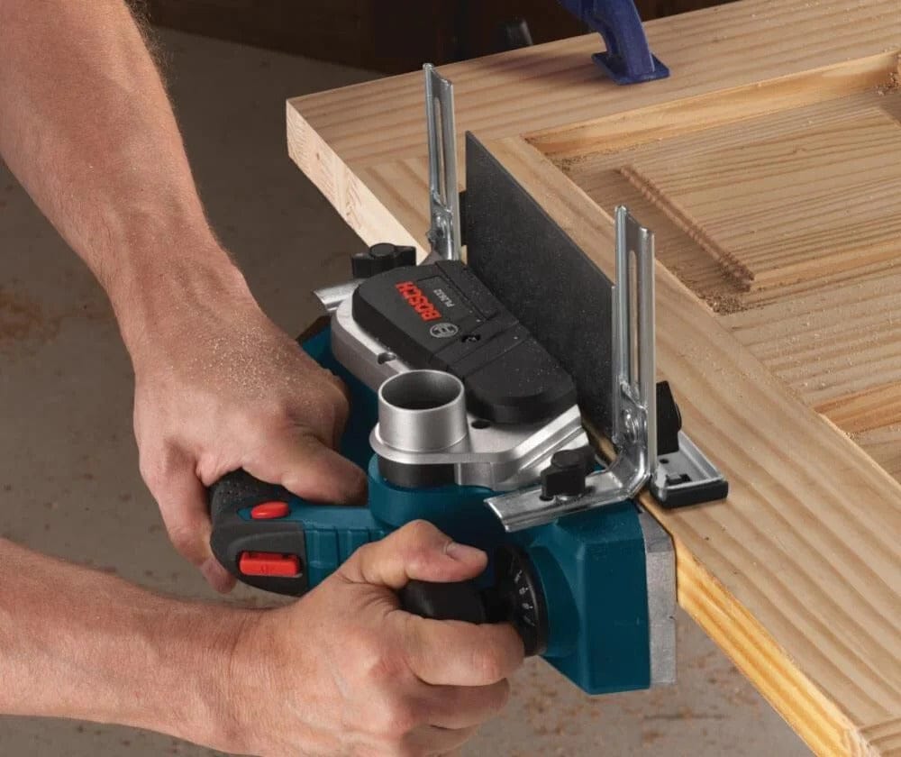 Bosch electric hand planer smoothing edge of clamped wooden board; guide fence ensures straight cut.