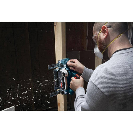 Person using power planer to shave vertical wooden beam; safety goggles and dust mask worn as wood shavings fly