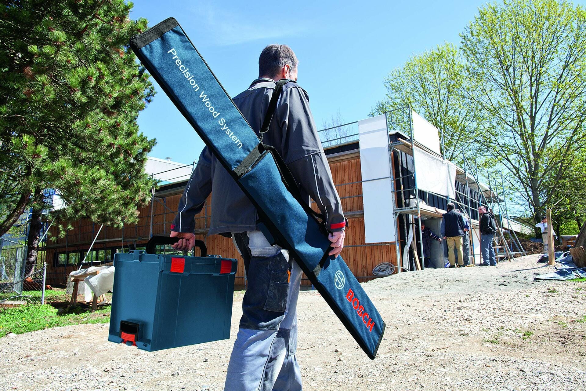 A construction worker carries the Bosch track saw guide rail kit in its blue bag slung over his shoulder while walking onto a jobsite.