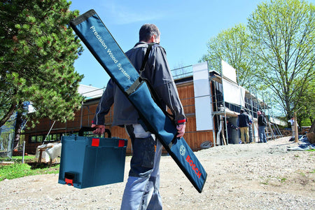 A construction worker carries the Bosch track saw guide rail kit in its blue bag slung over his shoulder while walking onto a jobsite.