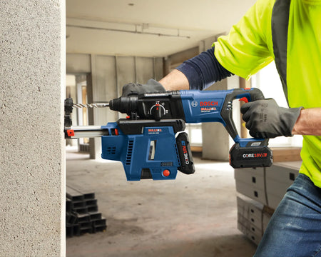A jobsite product image showing a worker in a high-visibility yellow t-shirt and safety gloves boring into a structural concrete pillar using a blue Bosch Bulldog 18V cordless rotary hammer drill equipped with a specialized onboard HEPA dust extraction attachment.