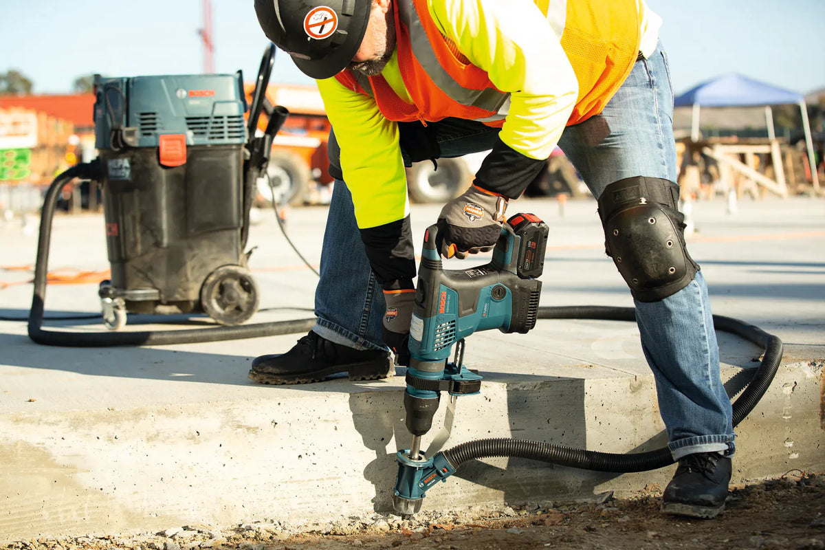 A low-angle construction photo showing a worker in safety gear, knee pads, and an orange safety vest drilling anchor holes vertically into a concrete foundation ledge with a Bosch hammer drill and vacuum shroud system connected to a heavy-duty portable dust extractor.