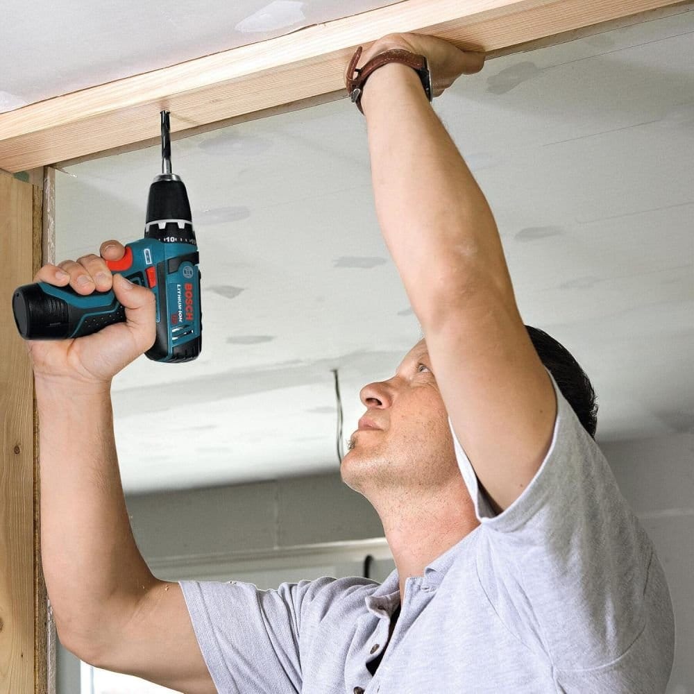 Action shot of a technician using a Bosch 12V cordless drill overhead to drive a screw into a wooden frame, demonstrating the battery's lightweight and balanced design.