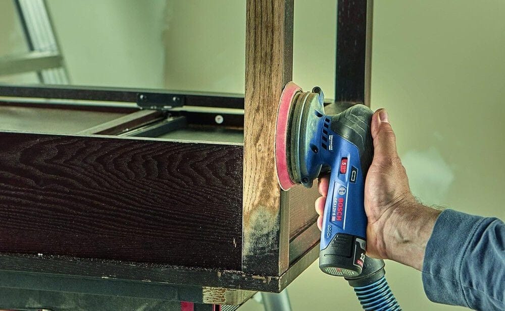 Close-up of a Bosch 12V cordless orbital sander being used on a dark wooden cabinet, powered by the compact 12V Max battery.