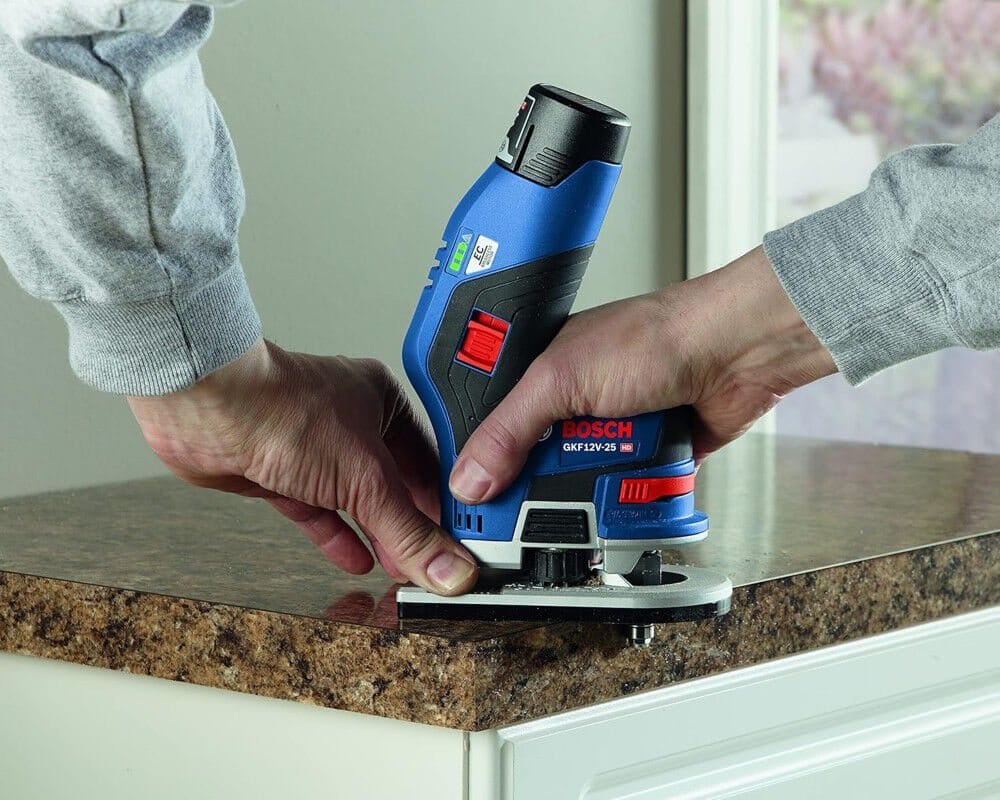 A person using a Bosch 12V cordless palm router on a countertop edge, with the BAT414 battery providing power in a small form factor.