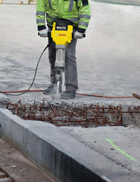 Person using Bosch BH2760VC Brute Breaker Hammer on concrete surface, gripping ergonomic handles during demolition work in a workshop setting.