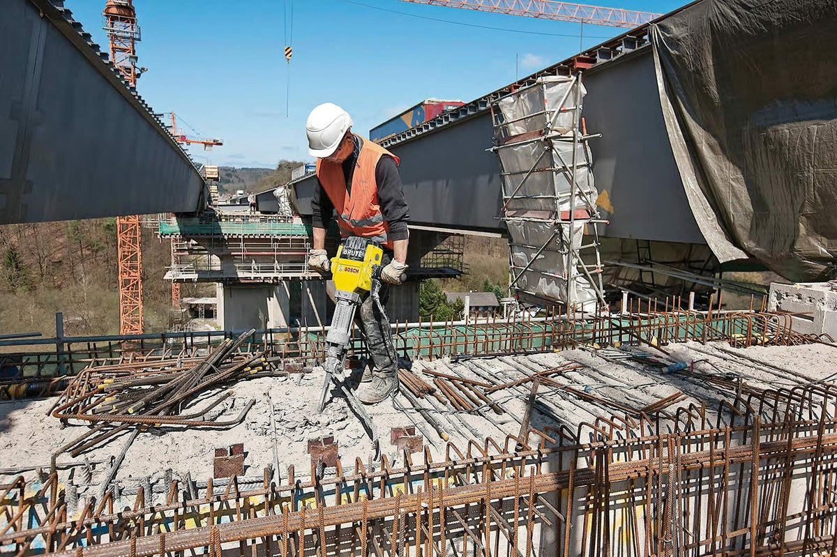 Person using Bosch BH2760VC Brute Breaker Hammer on concrete surface, gripping ergonomic handles during demolition work in a workshop setting.