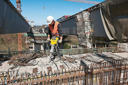 Person using Bosch BH2760VC Brute Breaker Hammer on concrete surface, gripping ergonomic handles during demolition work in a workshop setting.
