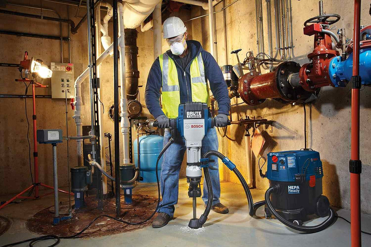 A construction worker wearing safety gear uses the Bosch BH2770VCD Brute Turbo 120V heavy-duty breaker hammer to break concrete flooring in an indoor utility basement, demonstrating the tool's compatibility with an attached Bosch dust extraction vacuum system.