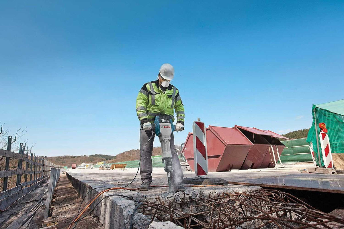 A wide outdoor construction scene showing a worker using the 63lb Bosch Brute Turbo breaker hammer to demolish a concrete slab and expose rebar on a major highway overpass project.