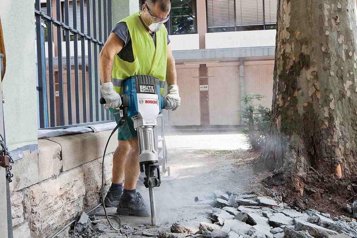 A construction worker uses the Bosch heavy-duty hex breaker hammer to break up an outdoor concrete sidewalk beside a building foundation and a large tree, showcasing its concrete removal power in tight outdoor spaces.