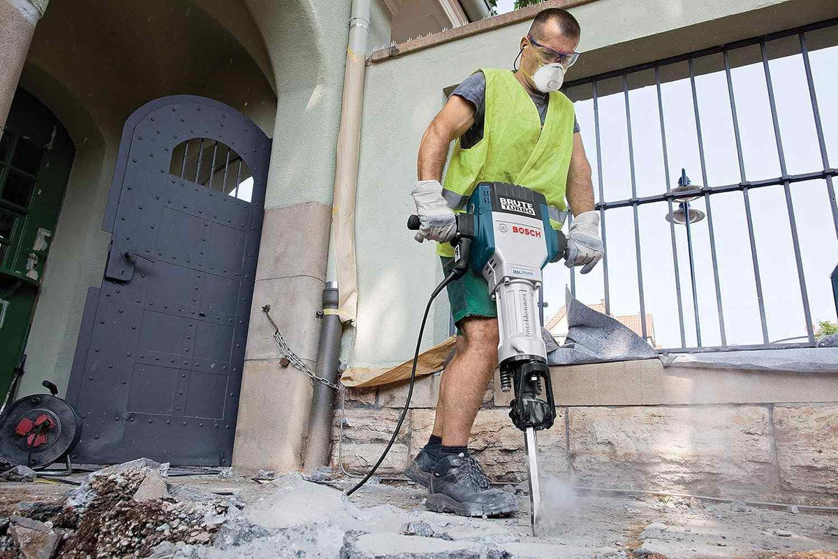 A worker wearing a dust mask and safety glasses operates the Bosch Brute Turbo 120V concrete demolition hammer to break up a thick concrete pathway directly outside a stone building with a metal door.