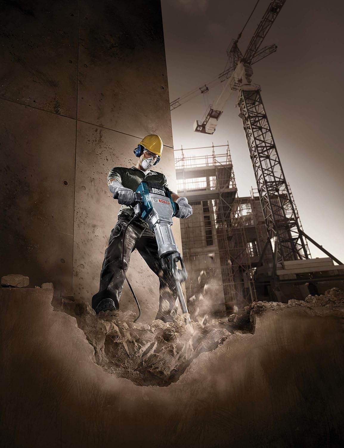 A dramatic low-angle action photo of a construction worker using the Bosch BH2770VCD Brute Turbo demolition breaker hammer to smash heavily through a thick concrete slab, with a large construction crane silhouetted in the background.
