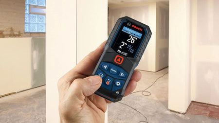 Action shot of a hand holding the GLM165-27C inside a room under construction, with the color display showing a measurement and the rounding function active