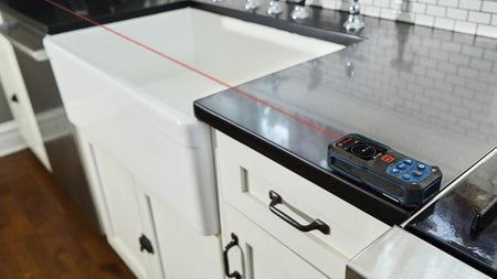 Action shot showing the Bosch BLAZE GLM165-27C laser measure sitting on a countertop, projecting the red laser beam across a kitchen sink to take an accurate indoor measurement