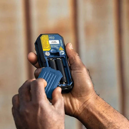 Close-up view of hands opening the battery compartment of the laser measure, showing the bay for inserting or replacing the two AA batteries