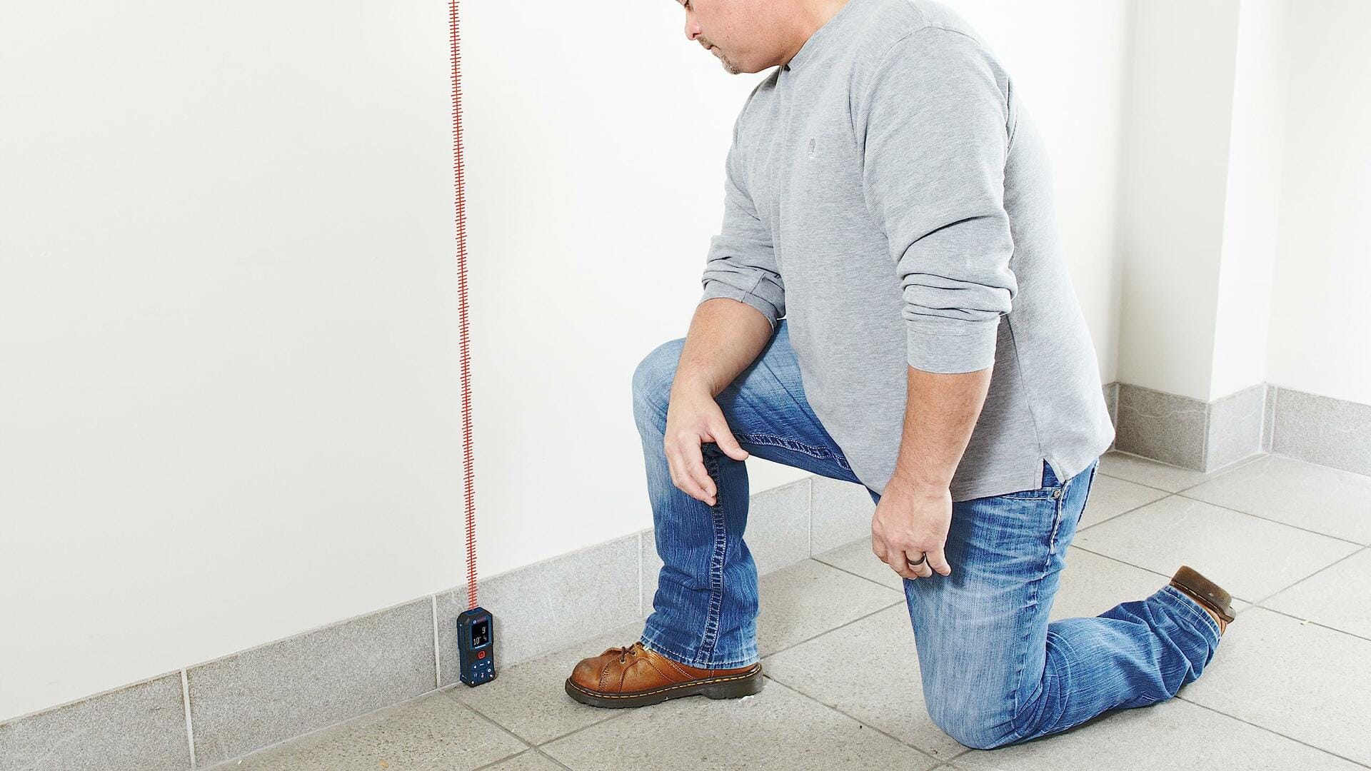 Man kneeling on tiled floor using Bosch GLM165-22 laser distance measure with red laser beam on white wall