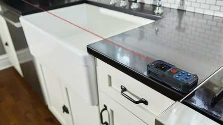BOSCH GLM165-22 laser distance measure in use on kitchen countertop measuring distance with red laser beam visible