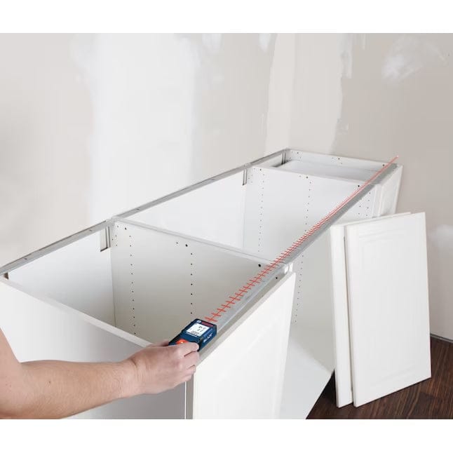 A hand holding the Bosch GLM165-10 laser distance measurer to size up the length of white kitchen cabinetry during an indoor installation project.