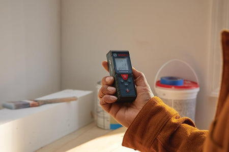 A professional contractor holding the Bosch BLAZE Pro laser distance measure in an indoor residential setting, displaying real-time distance and calculation readings on the backlit screen.
