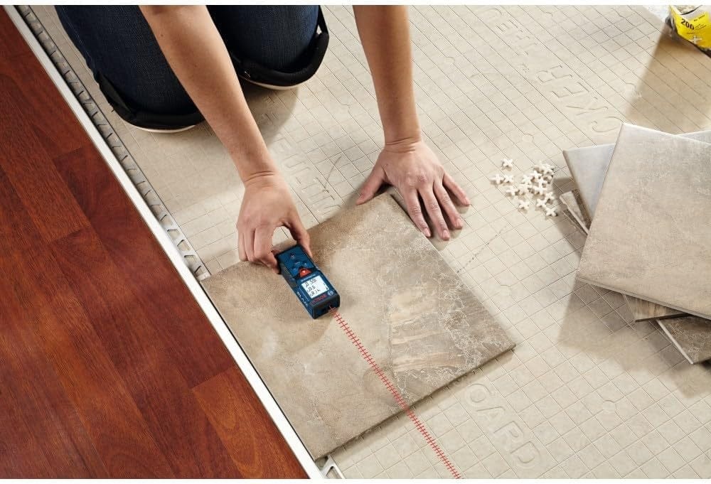 A professional installer on a jobsite using the Bosch BLAZE Pro laser measure to project a red laser beam across floor tiles to ensure precise layout and alignment.