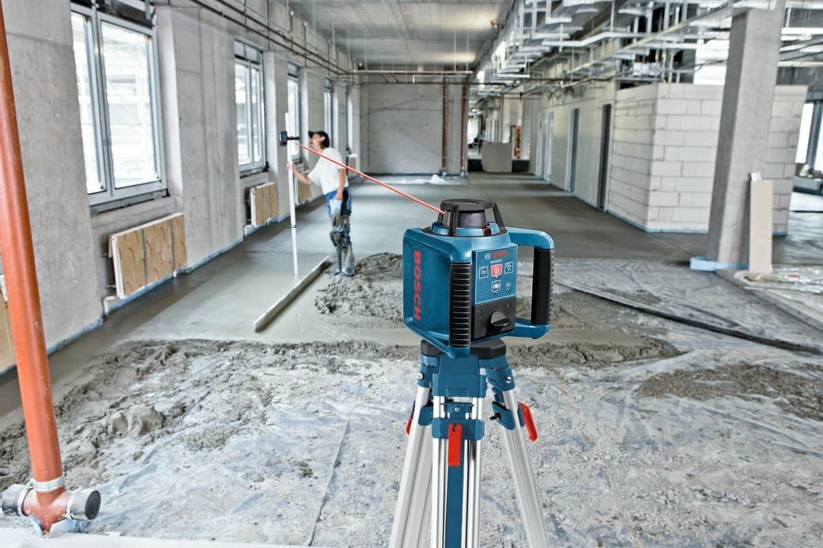 A real-world commercial construction application shot showing a blue Bosch automatic rotary laser level mounted securely onto the flat head of the BT160 aluminum tripod, projecting a visible red leveling reference beam across a concrete floor project while an on-site technician measures the grade in the background.