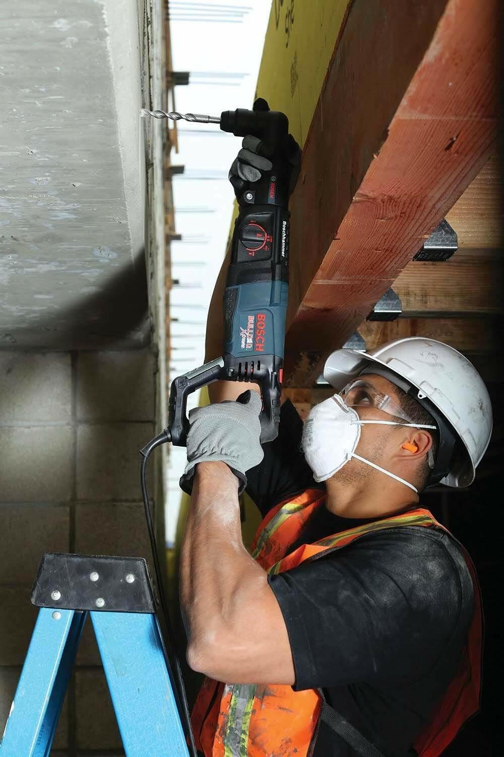 A construction worker in full safety gear uses the Bosch Bulldog rotary hammer to drill a hole upward into a concrete ceiling.