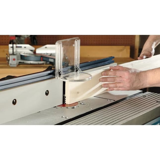 Action shot of a person using the Bosch RA1181 router table to rout the edge of a wooden board, with the clear plastic bit guard and tall fence visible for safety and support