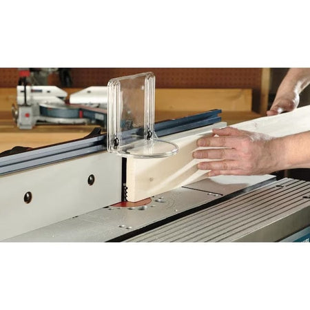 Action shot of a person using the Bosch RA1181 router table to rout the edge of a wooden board, with the clear plastic bit guard and tall fence visible for safety and support