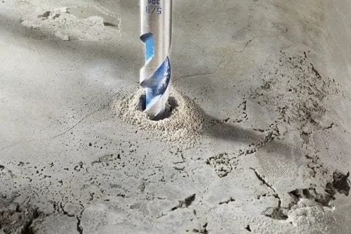 Close-up action shot of the drill bit penetrating a concrete surface, creating a pile of dust around the hole.