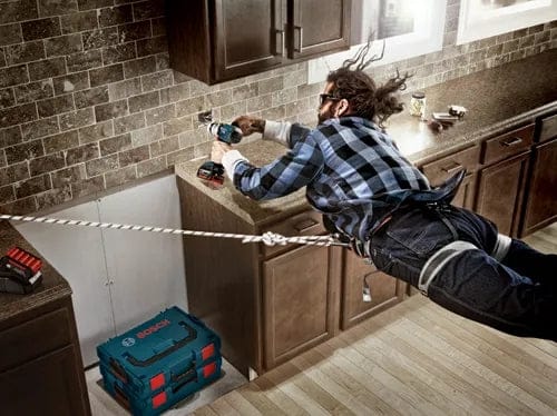 Humorous action shot of a worker suspended horizontally by ropes while drilling into a kitchen backsplash, emphasizing the "extreme" hold or power.
