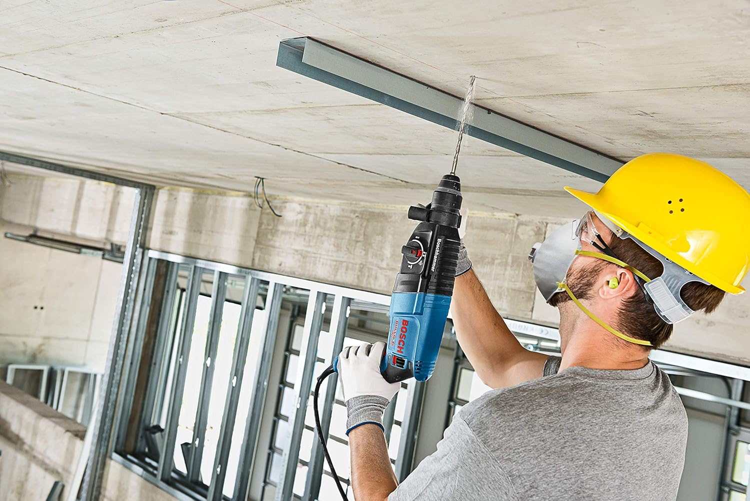 Low angle action shot of a construction worker wearing a yellow hard hat drilling overhead into a concrete ceiling.