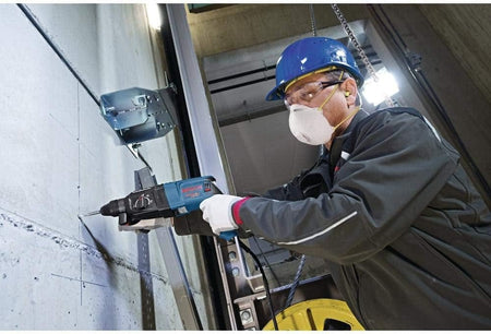 Action shot of a worker wearing a blue hard hat and safety glasses drilling an anchor hole into a concrete wall.