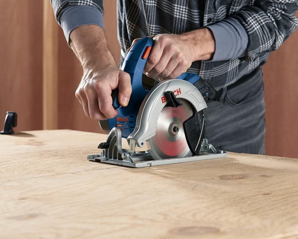 Person operating Bosch circular saw to cut thick wood plank; blade engaged and saw guided with both hands.