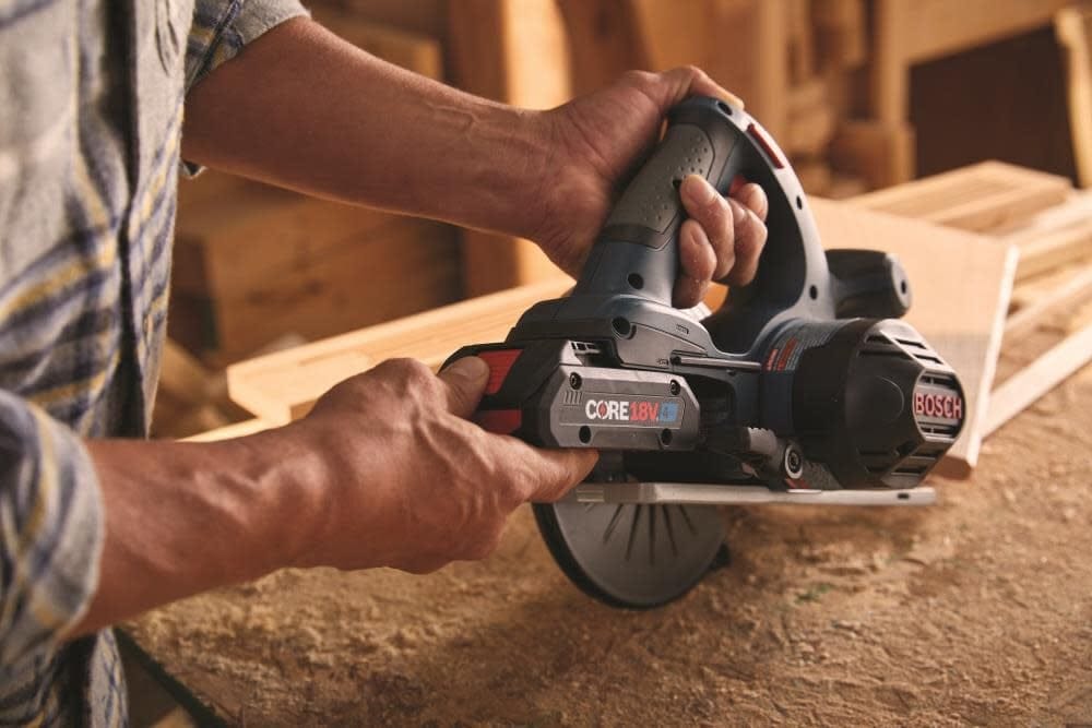 Person preparing Bosch CORE18V circular saw for use in workshop; wood boards and tools visible in background.