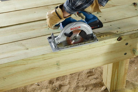 Person using Bosch cordless circular saw to cut wood on elevated beams; sawdust visible around active cutting area.