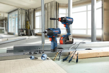 An indoor jobsite application lifestyle photo showing two blue Bosch cordless impact tools standing on a structural steel track framing plate inside a room under renovation, surrounded by loose jobsite accessories including black oxide twist drill bits, spade wood boring bits, nut drivers, and bi-metal hole saws.
