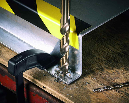 An action photograph of a cobalt drill bit being used to drill into a clamped metal workpiece, demonstrating the durability of the bit in a workshop setting.