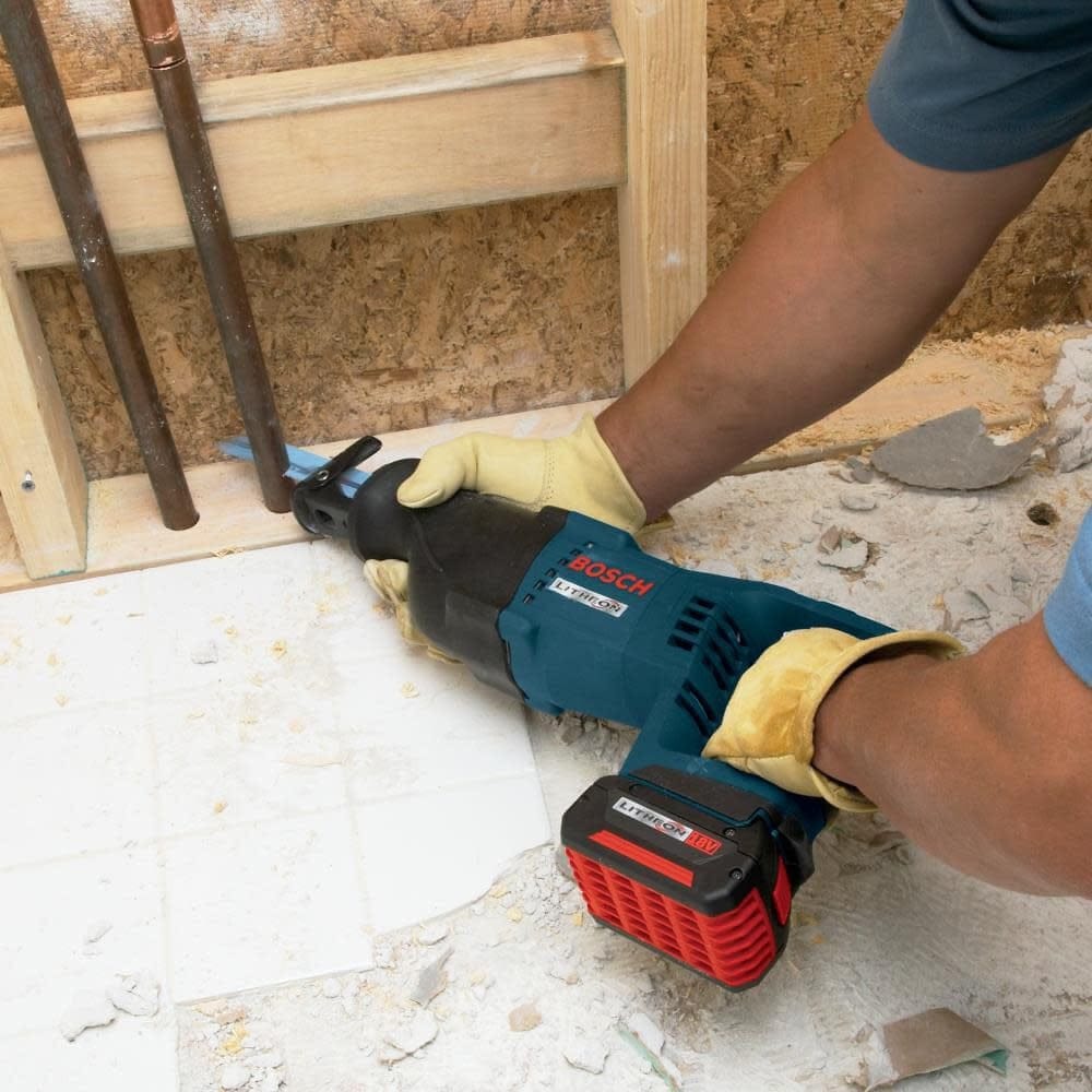 A person wearing leather work gloves using the Bosch reciprocating saw to cut through copper plumbing pipes near a wall plate.