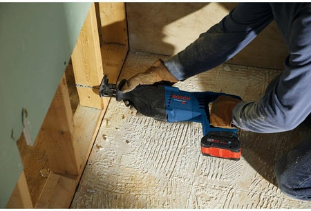 A construction worker wearing gloves using the Bosch reciprocating saw to cut through a wooden stud inside a wall cavity.