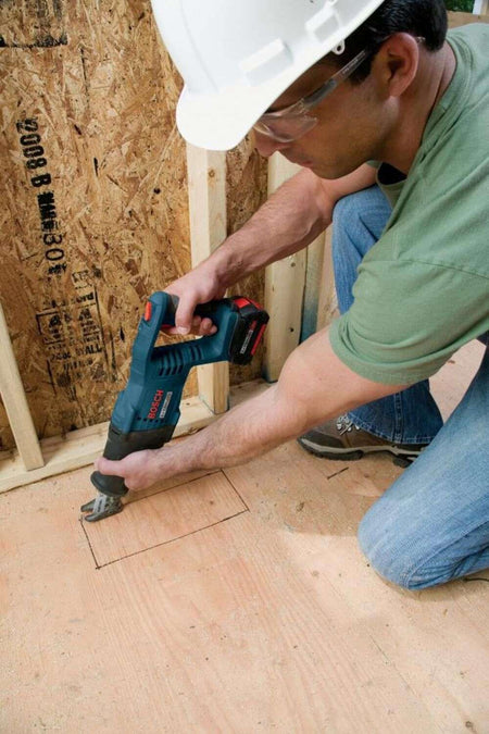 A construction worker wearing a hard hat kneeling on a plywood subfloor and using the reciprocating saw to cut a vent opening.