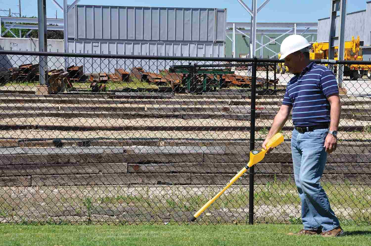 A worker wearing a white hard hat and safety gear uses the yellow magnetic locator to sweep a grass area next to a chain-link fence at an industrial job site.
