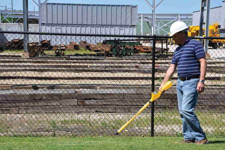 A worker wearing a white hard hat and safety gear uses the yellow magnetic locator to sweep a grass area next to a chain-link fence at an industrial job site.