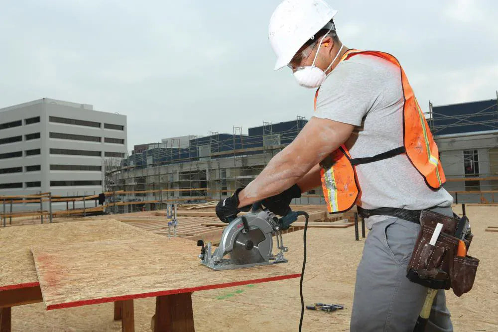An outdoor jobsite action photo of a construction carpenter wearing a white hard hat, safety glasses, safety vest, and dust mask as he uses the Bosch CSW41 15-amp worm-drive circular saw to make a rip cut through a sheet of oriented strand board OSB plywood resting on a sawhorse.