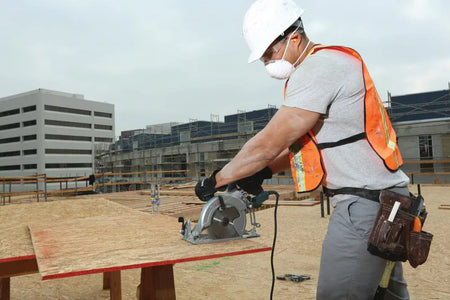 An outdoor jobsite action photo of a construction carpenter wearing a white hard hat, safety glasses, safety vest, and dust mask as he uses the Bosch CSW41 15-amp worm-drive circular saw to make a rip cut through a sheet of oriented strand board OSB plywood resting on a sawhorse.