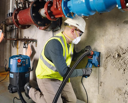 An action photograph showing a worker in safety gear using a Bosch angle grinder with a dust extraction shroud to cut a concrete wall near industrial pipes.