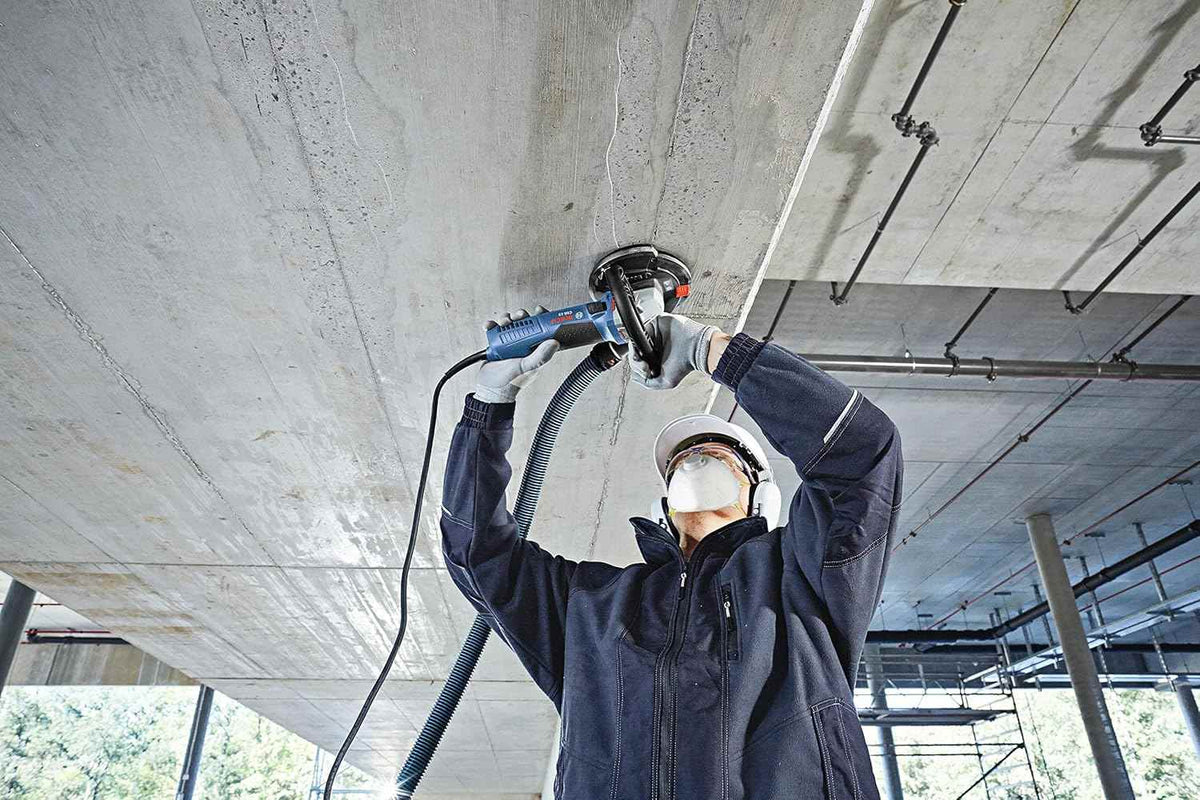 A close-up action shot of an operator on a scaffold using a Bosch grinder with a dust shroud to finish a concrete ceiling, highlighting the dust-free operation.